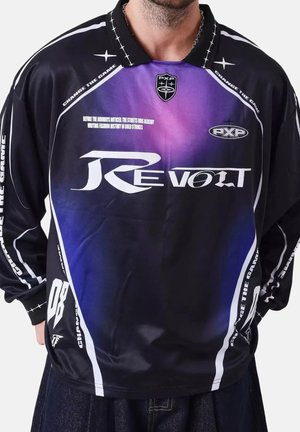 Homme portant un maillot de sport à manches longues noir et violet avec le texte blanc « Revolt », « Change the Game » et des logos sur le devant et les manches.