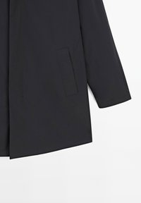Cappotto impermeabile nero con taglio dritto, dotato di tasca laterale e tessuto testurizzato. Le maniche sono leggermente svasate.