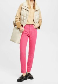 Jeans rosas de corte ajustado, chaleco acolchado beige, suéter claro y mocasines negros. Lleva un bolso tote beige claro.