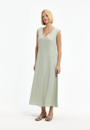 Woman with blonde bob haircut wearing a sleeveless, light green, V-neck maxi dress and snakeskin-patterned flat shoes standing on white background.