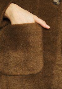 Hnědý vlněný kabát s měkkým, strukturovaným povrchem a velkou, čtvercovou kapsou, v níž spočívá ruka. Viditelný detail knoflíku na límci.