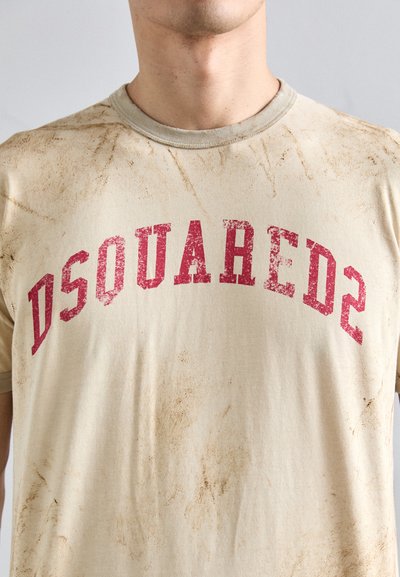 Beige Baumwoll-T-Shirt mit abgenutzter Textur, das einen verblassten roten "DSQUARED2"-Druck in gewölbter Schriftart zeigt. Sichtbare Schmutzflecken auf dem Stoff.