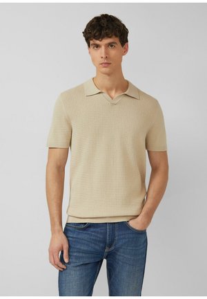 Mężczyzna ubrany w beżową, krótkorękawową, dzianinową koszulkę polo i niebieskie dżinsy, stojący z jedną ręką w kieszeni na jednolitym tle.