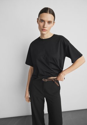 Femeie cu păr închis la culoare prins la spate poartă un tricou negru larg și pantaloni negri cu o curea maro, stând în fața unui fundal simplu, deschis la culoare.