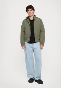 Oliven grønn polstret jakke med glidelås, svart polo skjorte, lyseblå vide jeans og svarte sko. Enkelt design, uten mønstre.