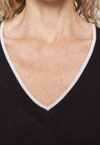 Zwarte trui met een v-hals en een witte rand. Het materiaal lijkt geribbeld en heeft een textuur rond de halslijn.