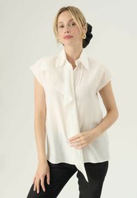 Blusa bianca a maniche corte con fronte arricciato, tessuto leggero, colletto e vestibilità rilassata, abbinata a jeans neri.