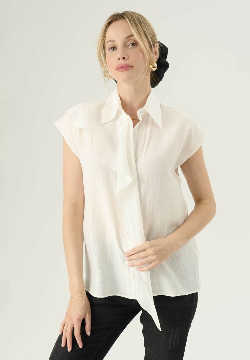 Blusa bianca a maniche corte con fronte arricciato, tessuto leggero, colletto e vestibilità rilassata, abbinata a jeans neri.