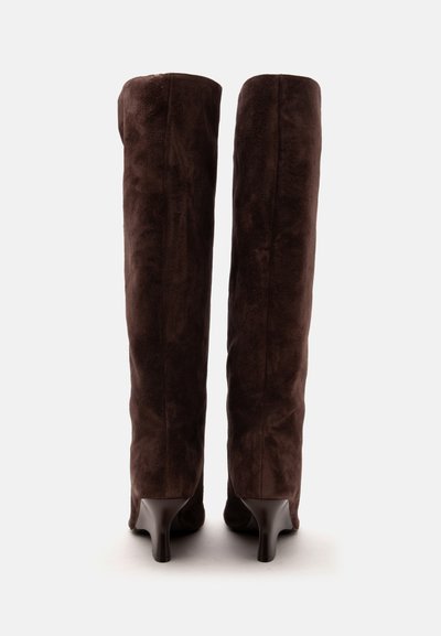 Paire de bottes hautes en suède marron foncé avec des talons fins, vues de dos sur un fond clair uni.