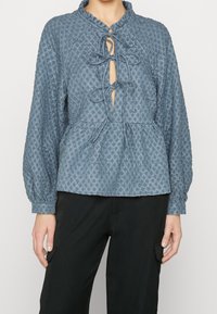 Blusa blu con texture, colletto, apertura a chiave e nastri. Caratterizzata da maniche lunghe e vita peplo, abbinata a pantaloni neri.