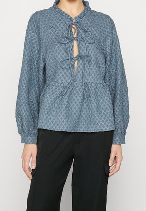 Blauwe blouse met textuur, kraag, sleutelgatopening en strikbandjes. Heeft lange mouwen en een peplum taille, gecombineerd met zwarte broeken.