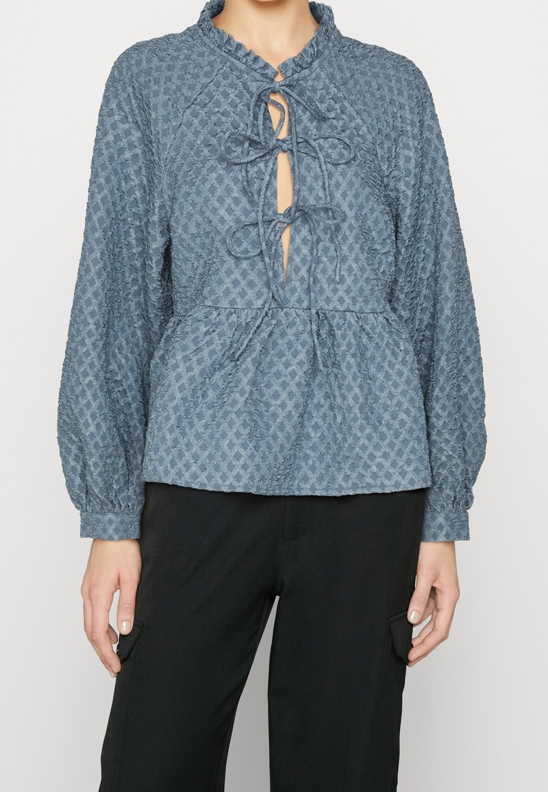 Blusa blu con texture, colletto, apertura a chiave e nastri. Caratterizzata da maniche lunghe e vita peplo, abbinata a pantaloni neri.