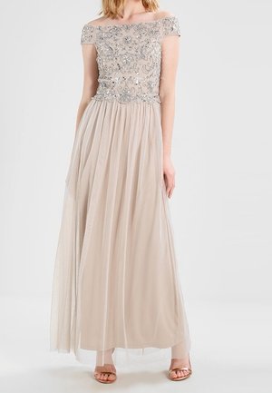 Femme portant une robe de soirée beige aux épaules dégagées, avec un corsage brodé de perles et de fleurs et une jupe en tulle fluide.