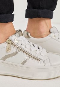 Fehér platform sportcipők fémes ezüst részletekkel, oldalsó cipzár és fűző, fekete felgöngyölt nadrággal, mezítláb boka körül.