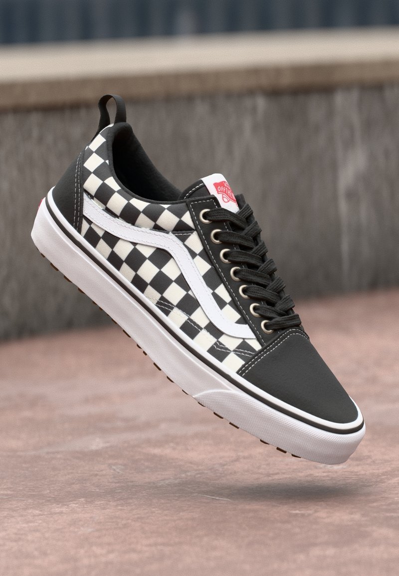 Baskets Vans à damier noir et blanc avec semelle blanche et lacets noirs, flottant au-dessus d'une surface en pierre.