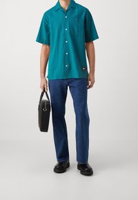 Camicia a maniche corte, di colore teal, con bottoni e un motivo testurizzato, abbinata a jeans blu e borsa nera. Presenta una tasca sul petto e un'etichetta con il logo.