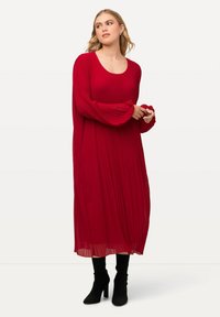 Rotes plissiertes Kleid mit langen Ärmeln, Rundhalsausschnitt und knöchellanger Silhouette, kombiniert mit schwarzen Stiefeletten mit Absatz. Glatter, fließender Stoff.