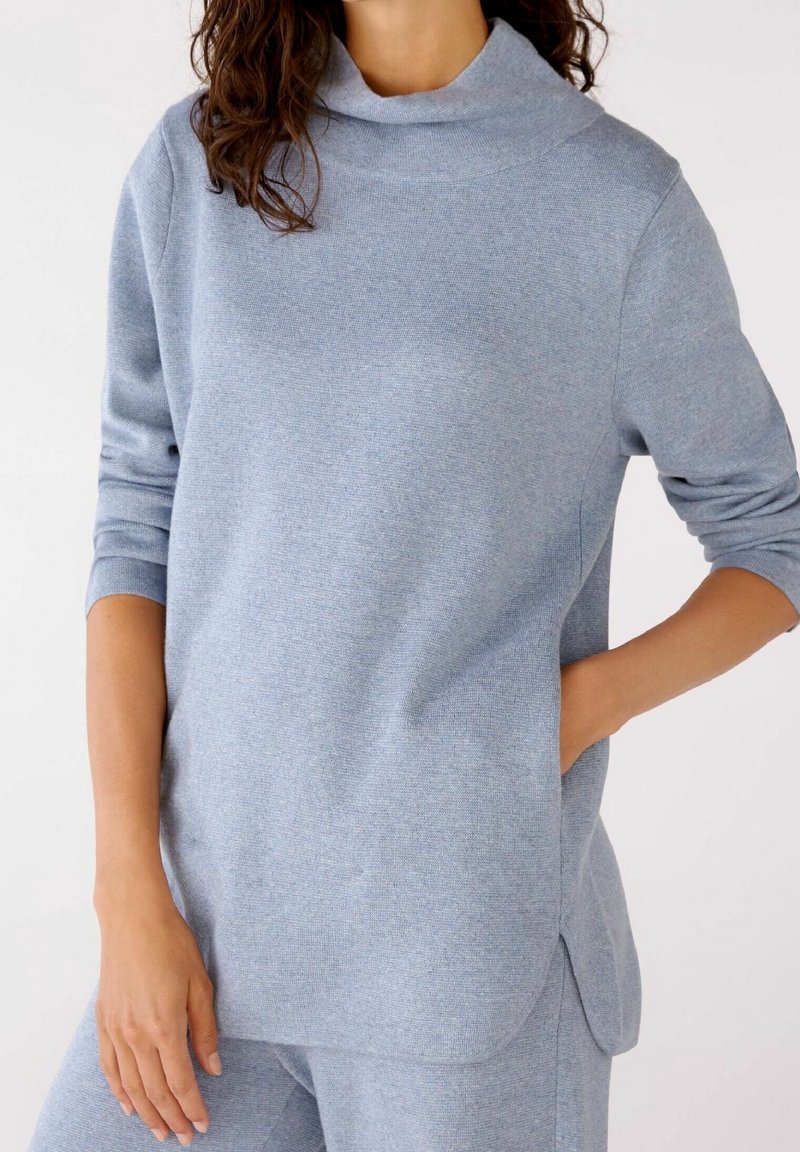 Femme portant un haut en maille bleu clair à manches longues et un pantalon assorti, une main dans la poche, sur un fond blanc uni.