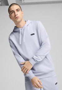 Lys grå hættetrøje med kengurulomme, der har et sort Puma-logo på brystet, lavet af et blødt og glat materiale.
