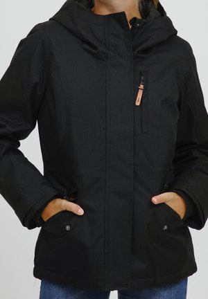 Personne portant une veste à capuche noire avec des poches zippées à l'avant et les mains dans les poches inférieures, vêtue d'un jean bleu.