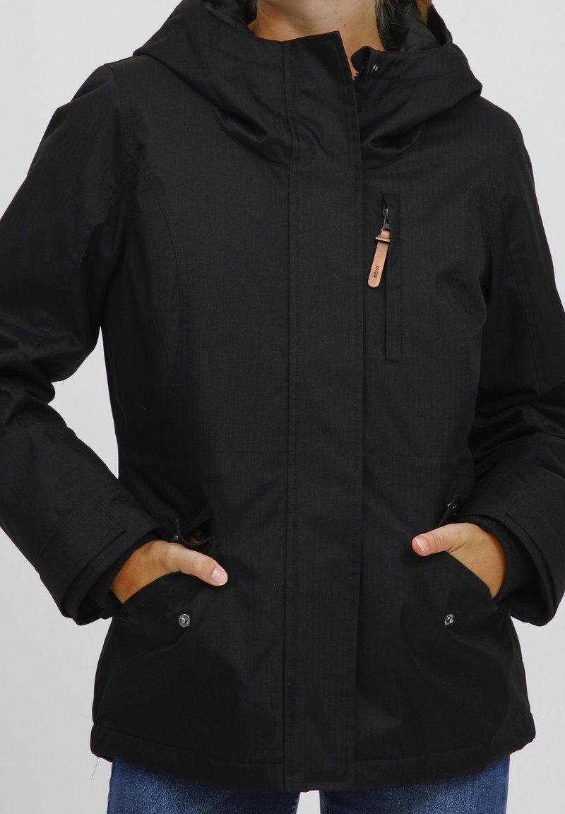 Personne portant une veste à capuche noire avec des poches zippées à l'avant et les mains dans les poches inférieures, vêtue d'un jean bleu.