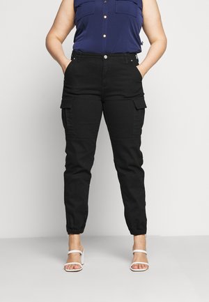 Osoba ubrana w czarne spodnie cargo z elastycznymi mankietami i granatową koszulę bez rękawów z guzikami, stojąca z rękami w kieszeniach w białych sandałach na obcasie.