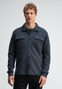 Grau-schwarzer Wollhemdjacke mit subtiler Textur, mit einem Kragen, zwei Brusttaschen und schwarzen Knöpfen, getragen über einem schwarzen T-Shirt.