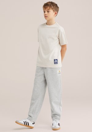 Dječak stoji s rukama iza leđa, nosi bež majicu kratkih rukava, sive trenirke s zakrpom i bijele tenisice s crnim prugama.