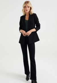 Svart skräddarsydd blazer med slagkrage, i kombination med en grön spets-topp och utsvängda svarta byxor. Slät tyg och välarbetad passform.