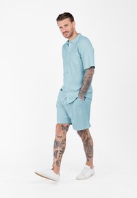 Hombre con tatuajes en las mangas y las piernas que lleva una camisa de botones de manga corta azul claro y pantalones cortos a juego, caminando con las manos en los bolsillos y zapatillas blancas.