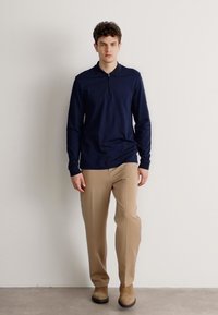 Camisa polo de manga longa em azul-marinho com colarinho com fecho, combinada com calças bege. A camisa apresenta um tecido texturizado e um corte descontraído.