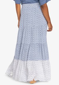 Mujer vestida con una larga falda de flores en tonos azul y blanco, con un top azul claro, de pie descalza frente a un fondo simple.