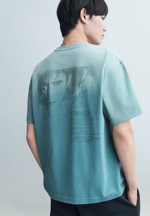 Νεαρός άνδρας που φοράει μεγάλης διάστασης t-shirt σε τιρκουάζ με ξεθωριασμένο μαύρο γραφικό ενός προσώπου και κείμενο στην πλάτη, στέκεται με το ένα χέρι στην τσέπη.