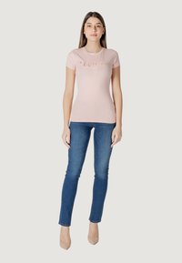 Camiseta ajustada de color rosa claro con un logotipo estilizado, combinada con jeans ajustados azules y tacones nude. Textura suave, diseño sencillo.