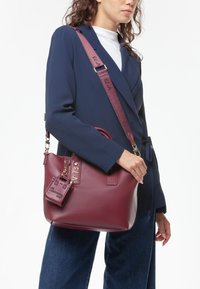 Bolso de cuero burdeos con forma estructurada, acentos en metal dorado y una etiqueta desmontable. Llevado con un blazer en contraste de color azul marino.