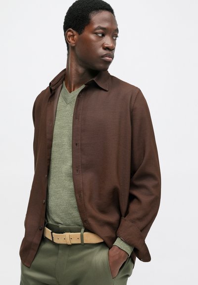 Homem a usar uma camisa castanha desabotada sobre um suéter verde em forma de V, calças verdes e um cinto bege, olhando para o lado com uma mão no bolso.