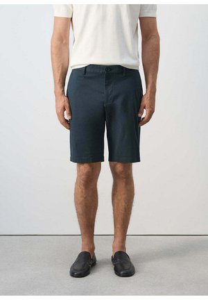 Hombre vistiendo pantalones cortos azul oscuro hasta la rodilla, camisa blanca de manga corta y zapatos negros sin cordones, de pie frente a un fondo sencillo.