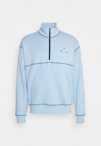 Světle modrá mikina s vysokým límcem, polovičním zipem a přední kapsou; obsahuje kontrastní prošívání a malé logo na hrudi.