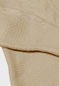 Beige gebreid sweaterstof heeft een zachte, gestructureerde oppervlakte met verhoogde patronen. Het materiaal lijkt warm en gezellig, geschikt voor koudere weersomstandigheden.