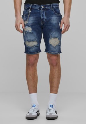 Denim kratke hlače u tamnoplavoj boji s poderanim rubovima i velikim distorziranih mrljama. Imaju lanac kao dodatak i uski kroj. Nosite ih s tenisicama.