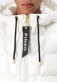 Giacca piumino bianca con finitura lucida, cordini neri, hardware dorato e un'etichetta del marchio visibile con la scritta "PINKO" stampata in bianco.