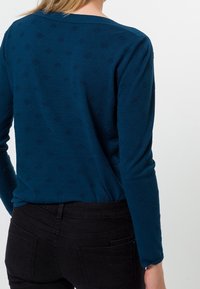 Donker teal textuurtrui met lange mouwen en een brede halslijn. Heeft subtiele verhoogde patronen en gecombineerd met zwarte aangesloten broek.