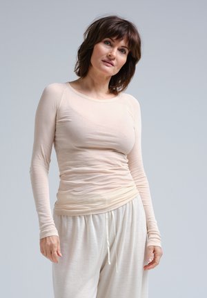 Beige Langarmoberteil mit Rundhalsausschnitt, leicht tailliertem Design, aus weichem, leichtem Material. Kombiniert mit lässigen weißen Hosen.