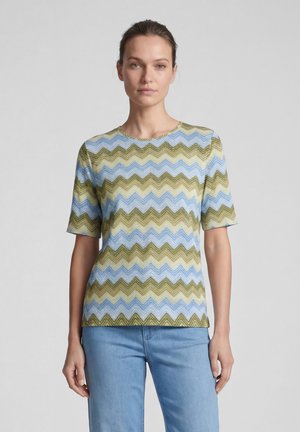 Frau mit zurückgebundenen Haaren, die ein kurzärmeliges, blau-grün gemustertes Zickzack-Shirt und hellblaue Jeans trägt, steht vor einem einfarbigen Hintergrund.