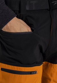 Schwarz-orange Hosen besitzen eine enge Passform mit einem strukturierten Bund. Eine mit Reißverschluss verschlossene Tasche befindet sich am Oberschenkel für sicheren Stauraum.