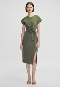 Olivgrünes Kleid mit kurzen Ärmeln, Knoten-Detail vorne und seitlichem Schlitz. Glatter, leichter Stoff, knielanges Design und minimalistische Ästhetik.
