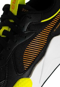 Scarpa sportiva nera e gialla con tomaia testurizzata, dettagli a strisce arancioni, collarino imbottito e intersuola bianca con suola esterna texturizzata.