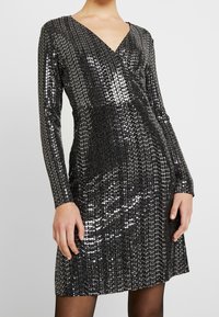 Skinande svart paljettklänning med långa ärmar, V-ringning och en figurnära överdel som smalnar av till en utsvängd kjol vid mitten av låret.