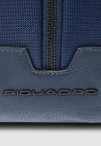 Sac en nylon bleu et cuir avec une finition texturée, une fermeture éclair noire centrale et le logo "Piquadro" embossé à la base.