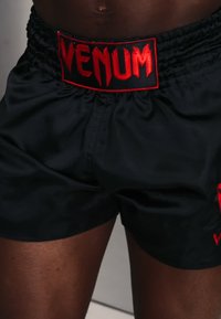 Pantalones cortos deportivos negros de la marca Venum, usados por una persona, mostrando la cintura elástica y el logo en letra roja.